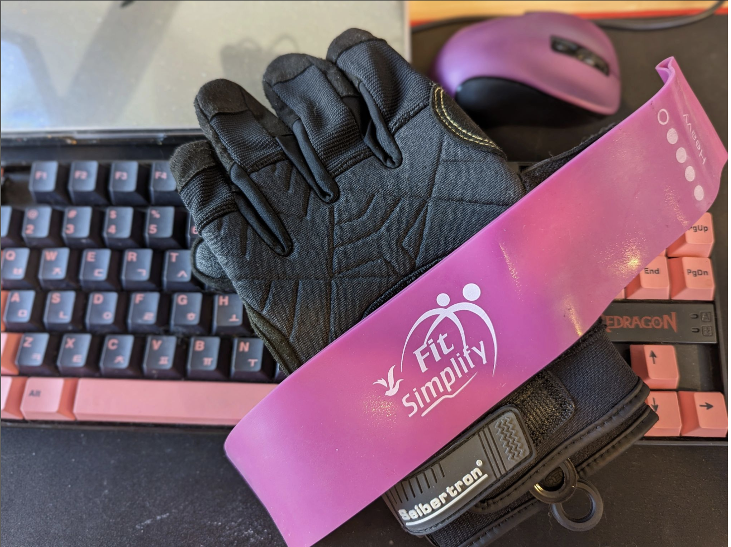 A purple stretch band and black workout gloves laying on a pink and black computer keyboard.